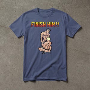 May include: A slate blue short-sleeve t-shirt featuring a pixel art graphic of two figures in a wrestling pose. Above the graphic, the text FINISH HIM!! is printed in a yellow and red gradient pixelated font. The shirt has a crew neckline.