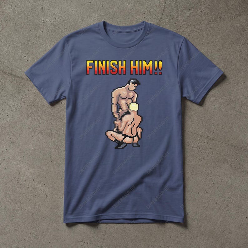 May include: A slate blue short-sleeve t-shirt featuring a pixel art graphic of two figures in a wrestling pose. Above the graphic, the text FINISH HIM!! is printed in a yellow and red gradient pixelated font. The shirt has a crew neckline.