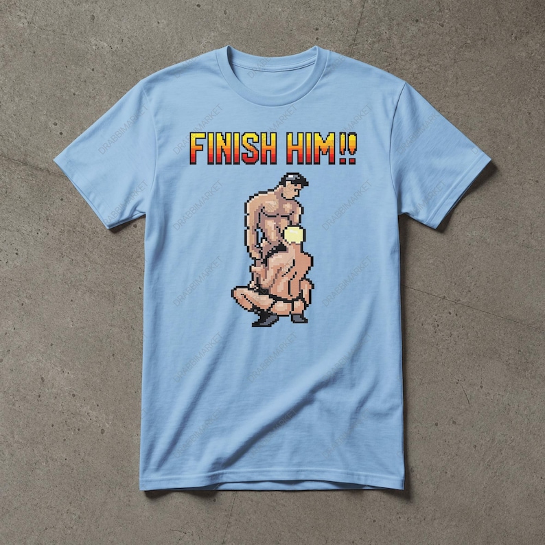 May include: A light blue short-sleeved t-shirt featuring a pixel art graphic of two figures in a wrestling pose. Above the graphic, the text FINISH HIM!! is printed in a yellow and red gradient font. The design is centred on the chest area.