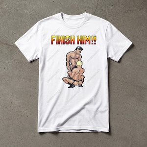 May include: A white short-sleeved t-shirt featuring a pixel art graphic of two figures in a sexual position. Above the graphic, the text FINISH HIM!! is printed in a yellow and red pixelated font. The shirt has a crew neckline and short sleeves.