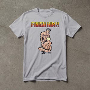 May include: A light grey short-sleeved t-shirt featuring a pixel art graphic of two figures in a combat stance. Above the figures, the text FINISH HIM!! is printed in a yellow and red gradient font with a black outline on the chest area.