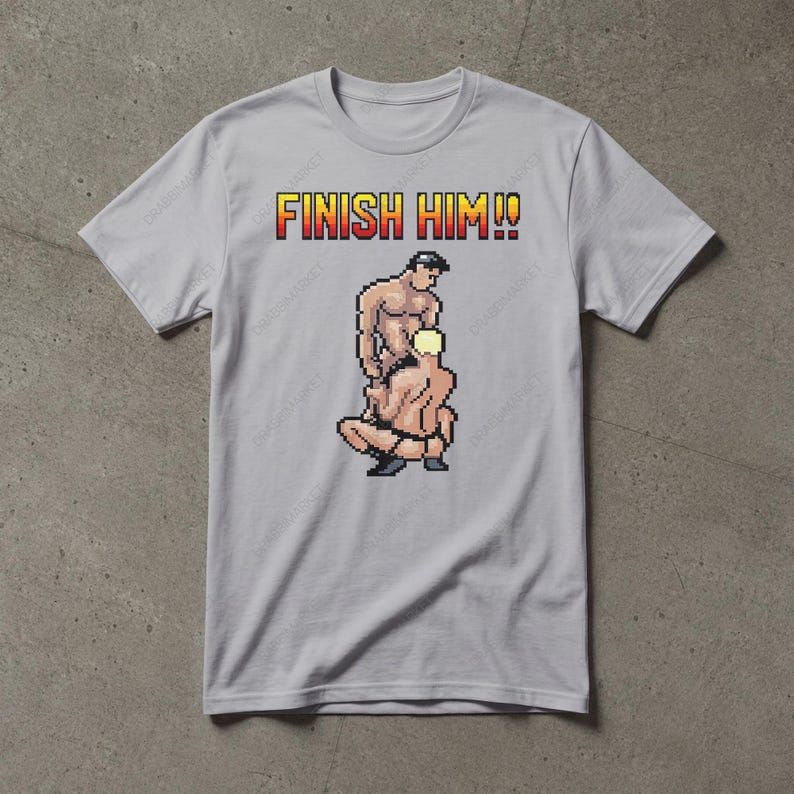 May include: A light grey short-sleeved t-shirt featuring a pixel art graphic of two figures in a combat stance. Above the figures, the text FINISH HIM!! is printed in a yellow and red gradient font with a black outline on the chest area.