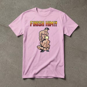 May include: A light pink short-sleeved t-shirt featuring a pixel art graphic of two figures in a wrestling pose. Above the figures, the text FINISH HIM!! is printed in a bold, yellow and red pixelated font. The shirt has a crew neckline.