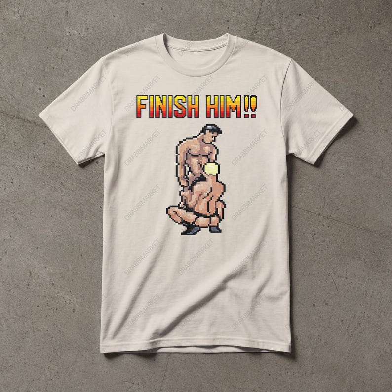 May include: A beige short-sleeved t-shirt featuring a pixel art graphic of two figures in a sexual position with the text FINISH HIM!! in a red and yellow gradient font above the illustration. The shirt has a crew neckline and a standard fit.