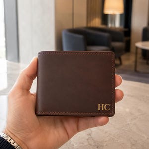 Può includere: Un portafoglio bifold in pelle marrone scuro con una finitura opaca liscia e cuciture visibili lungo i bordi. L'angolo in basso a destra del portafoglio è impresso con le lettere maiuscole dorate HC in un classico carattere serif.
