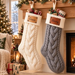 Op de afbeelding: Twee gebreide kerstsokken met kabelpatroon hangen aan een houten schoorsteenmantel. De ene sok is crèmekleurig met een leren label met de naam Grace, de andere is grijs met de naam Henry. Beide hebben witte geribbelde boorden en lussen.