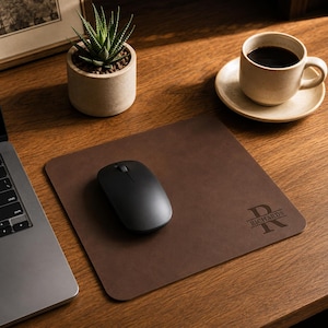 May include: A square dark brown leather mouse pad featuring a personalized monogram design with a large letter R and the name RICHARDS printed in a clean font. The mouse pad has rounded corners and a smooth surface, with a black computer mouse placed on top.