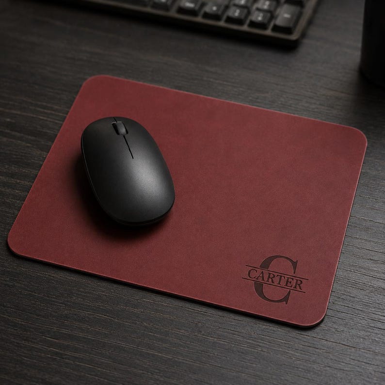 May include: A rectangular dark red leather mouse pad with rounded corners. The surface features a black engraved monogram consisting of a large letter C with the name CARTER centered horizontally across it. A black wireless computer mouse sits on top.