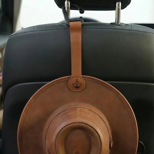 Könnte beinhalten: Ein brauner Lederhut, der an einer Kopfstütze im Auto hängt. Der Hut wird von einem braunen Lederriemen mit einem Metallhaken gehalten. Der Riemen hat ein rundes Emblem mit einem Anker und den Buchstaben "BC".