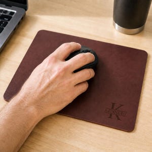 May include: A rectangular dark brown leather mouse pad featuring a personalized engraving in the bottom right corner. The engraving displays a large letter K with the name KENNEDY centered horizontally across the letter in a serif font.