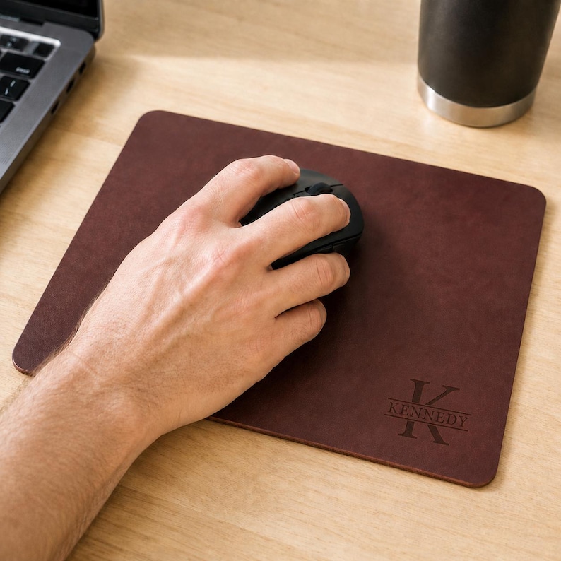 May include: A rectangular dark brown leather mouse pad featuring a personalized engraving in the bottom right corner. The engraving displays a large letter K with the name KENNEDY centered horizontally across the letter in a serif font.