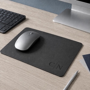 May include: A rectangular black mouse pad with rounded corners featuring the embossed letters C|N in the bottom right corner. A smooth grey wireless computer mouse rests on the left side of the pad, which is positioned on a light wood grain surface.