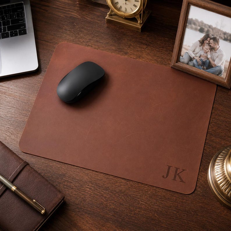 May include: A rectangular brown leather desk mat featuring rounded corners and the embossed initials JK in the bottom right corner. The smooth surface of the mat provides a workspace for a black wireless computer mouse placed on the left side.