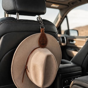 May include: A tan felt hat with a brown leather band hanging from a brown leather hat clip. The clip features a teardrop shape with a dark brown anchor graphic and the letters B and C engraved on it, attached to a metal hook on a car headrest.
