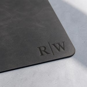 May include: A rectangular dark gray desk mat with a smooth, matte surface. The bottom right corner features a debossed monogram consisting of the capital letters R and W separated by a vertical line, creating a clean and professional appearance.