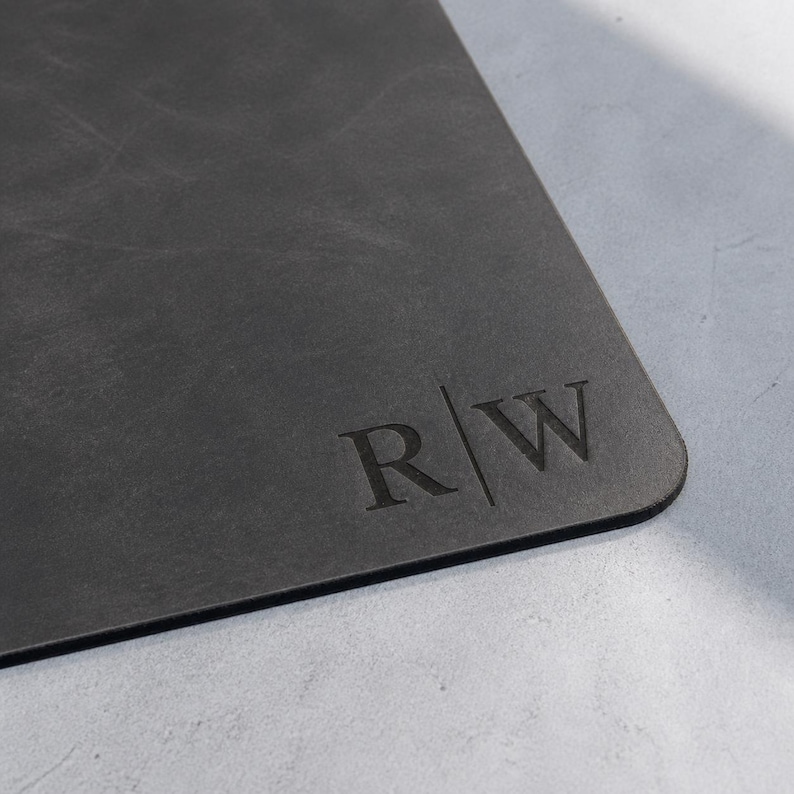 May include: A rectangular dark gray desk mat with a smooth, matte surface. The bottom right corner features a debossed monogram consisting of the capital letters R and W separated by a vertical line, creating a clean and professional appearance.