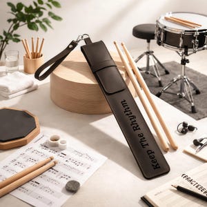 Puede incluir: Un estuche de cuero marrón con la frase "Keep The Rhythm" y un par de baquetas de madera. También se ven una batería, una almohadilla de práctica y partituras.