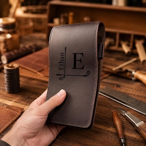 Peut inclure: Un porte-carte de score de golf en cuir marron foncé avec un motif gravé au laser comprenant le nom Ethan, la lettre E et deux icônes de clubs de golf. L'accessoire rectangulaire présente des coutures visibles et une finition mate lisse.