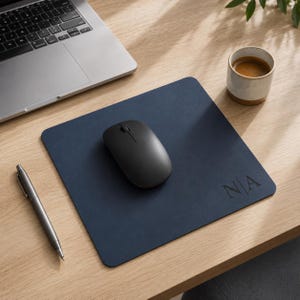May include: A square navy blue leather mouse pad featuring the embossed letters N|A in the bottom right corner. The mouse pad has a smooth surface and rounded corners, with a black wireless computer mouse placed in the center of the pad.