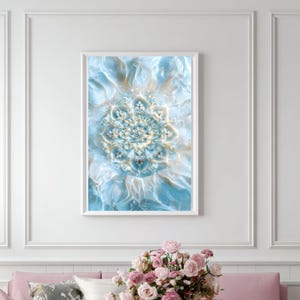 May include: Framed wall art featuring a detailed, abstract floral design. The artwork showcases a central, symmetrical pattern in shades of light blue, white, and gold, set against a soft, flowing background. The piece is encased in a white frame.