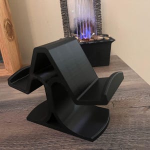May include: A black, 3D-printed phone stand. The stand has a unique, curved design with two cradles to hold a phone. The stand is made of a matte black material and sits on a wooden surface.