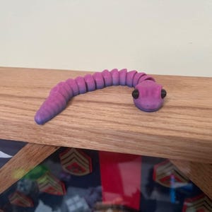 May include: A segmented, articulated toy snake in shades of purple and blue. The snake has black eyes and is resting on a wooden surface. The toy is designed to be flexible and poseable.