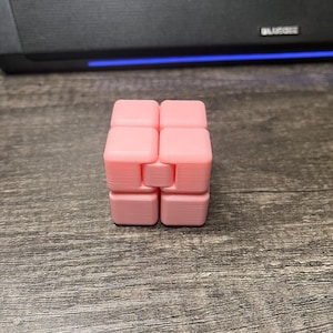 Può includere: Un cubo fidget rosa dal design unico. Il cubo è composto da otto blocchi quadrati collegati tra loro. I blocchi sono di colore rosa chiaro uniforme. Il cubo è appoggiato su una superficie di legno.