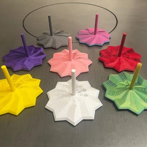 May include: A collection of eight colorful, star-shaped 3D-printed objects. Each has a central, vertical post. Colors include yellow, white, green, red, pink, purple, and gray. The objects are arranged on a gray surface.