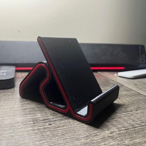 Può includere: Un supporto nero per telefono con bordi rossi. Il supporto è progettato per tenere un telefono o un tablet in posizione verticale. È realizzato in un materiale robusto e ha un design elegante e moderno.