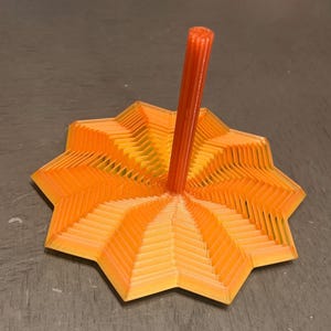 May include: A 3D-printed orange and yellow star-shaped toy with a central orange rod. The toy has a layered design, with the star's points radiating outwards. The rod is cylindrical and stands upright, adding a vertical element to the design.