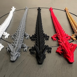 May include: Five 3D-printed dragon figurines in a row. The dragons are white, gray, black, red, and gold. Each dragon has a spiky back and detailed legs. The figurines are lying on a dark surface.