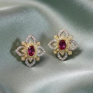 May include: A pair of floral-shaped stud earrings. Each earring features a central oval ruby gemstone, surrounded by a gold and silver filigree design. The earrings are set against a soft, light green background.