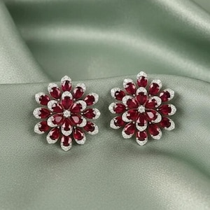 Op de afbeelding: Een paar bloemvormige oorstekers. Elke oorbel heeft een cluster van dieprode edelstenen omgeven door een halo van kleinere, heldere stenen. De oorbellen zijn geplaatst tegen een zachte, saliegroene achtergrond.