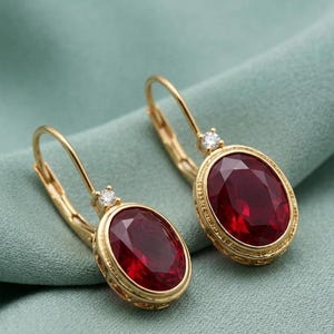 May include: A pair of gold-colored earrings with oval, deep red gemstones. Each earring features a small, clear accent stone and a decorative gold border. The earrings are set against a soft, teal-green background.