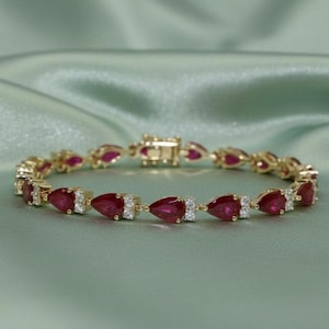 以下が含まれることがあります： ゴールドのブレスレット。涙型の深い赤色の宝石が特徴です。各宝石は小さな透明な石で装飾されています。ブレスレットは柔らかい緑色の背景に配置され、ジュエリーの輝きとデザインを際立たせています。