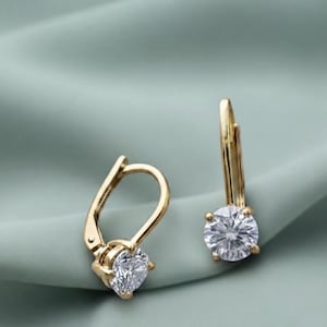 May include: A pair of gold leverback earrings, each featuring a single round, clear, faceted gemstone held in a four-prong setting. The metal has a polished finish, and the design includes a hinged mechanism for secure closure on the ear.