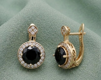 May include: A pair of gold-toned earrings featuring a large, round black stone surrounded by a halo of small, clear stones. The earrings have a lever back closure and a small cluster of stones at the top.