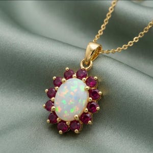 May include: A gold pendant necklace featuring an oval opal gemstone surrounded by small, round, red gemstones. The opal has iridescent colors. The necklace chain is also gold.