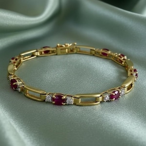 Könnte beinhalten: Ein goldenes Armband mit abwechselnd oval geschliffenen roten Edelsteinen und kleinen, runden, klaren Edelsteinen. Das Armband hat ein rechteckiges Kettendesign und einen Verschluss. Der Schmuck wird auf einer hellgrünen Satin-Oberfläche präsentiert.