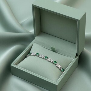 May include: A silver bracelet with emerald and diamond accents rests inside a sage green jewelry box. The box is open, revealing the bracelet against a soft, light green interior. The background is a silky, light green fabric.