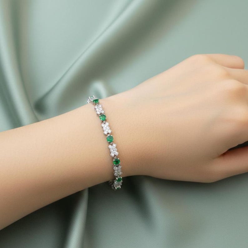 May include: A silver bracelet with alternating emerald green and clear, sparkling stones. The bracelet is displayed on a person's wrist against a soft, light green background. The bracelet's design features a repeating pattern of the gemstones.