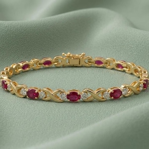 May include: A gold-toned bracelet with alternating oval-cut red gemstones and small, round, clear gemstones. The bracelet features an X-shaped design between the gemstones and a clasp closure. The background is a solid, muted green.