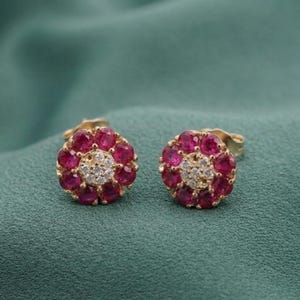 May include: A pair of gold-colored stud earrings. Each earring features a floral design with a cluster of small, round, deep red gemstones surrounding a center of smaller, clear gemstones. The earrings are set against a teal green background.