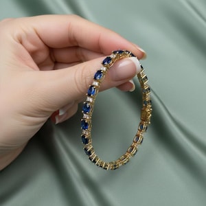 Puede incluir: Una pulsera dorada con piedras preciosas ovales de color azul oscuro y pequeñas piedras claras. La pulsera se muestra sobre un fondo verde suave, destacando su elegante diseño. Una joya clásica.