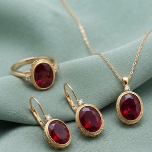 May include: A gold jewelry set consisting of a ring, a pair of lever-back earrings, and a pendant necklace. Each piece features a central oval-cut deep red gemstone surrounded by a gold frame, accented with a single small clear stone at the top.