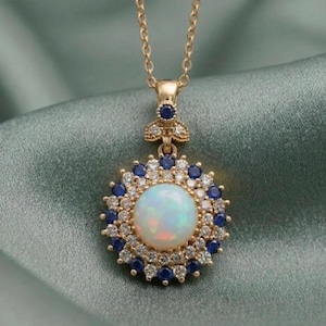Pode incluir: Um colar com pendente em ouro apresentando um cabochão de opala redondo central rodeado por um halo duplo de pequenos diamantes brancos e safiras azuis. O pendente pende de uma corrente de ouro fina, com uma safira azul na argola.