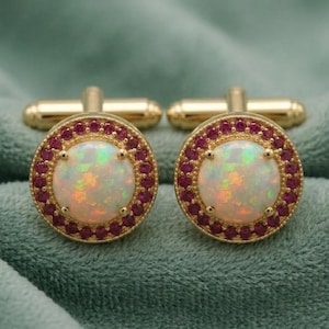 May include: A pair of gold cufflinks. Each cufflink has a round, iridescent opal centre stone encircled by small, red gemstones. The cufflinks have a classic, elegant design, suitable for formal wear.