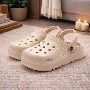 Könnte beinhalten: Ein Paar beigefarbene Plateau-Clogs mit mehreren Belüftungslöchern. Die Clogs haben eine dicke, geriffelte Sohle und einen verstellbaren Riemen. Die Schuhe sind aus einem glatten, einfarbigen Material.