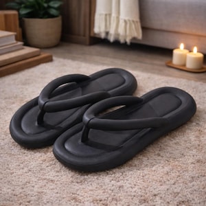 Peut inclure: Une paire de tongs noires avec des semelles épaisses et un montant arrondi. Les sandales sont faites d'un matériau souple et sont conçues pour le confort. Elles sont posées sur un tapis beige.
