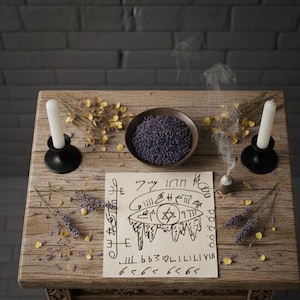 Pode incluir: Uma mesa de madeira rústica exibe uma cena ritualística. Duas velas brancas em suportes pretos ladeiam uma tigela de lavanda roxa e um pergaminho com símbolos. Fumaça de incenso sobe, com flores secas espalhadas.